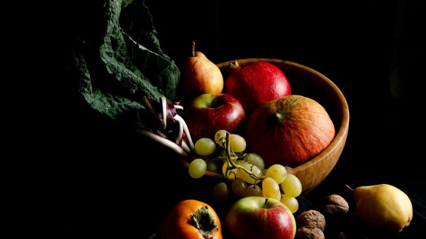 Dark still life composition featuring assorted autumn fruits in a wooden bowl, creating a moody and artistic scene.