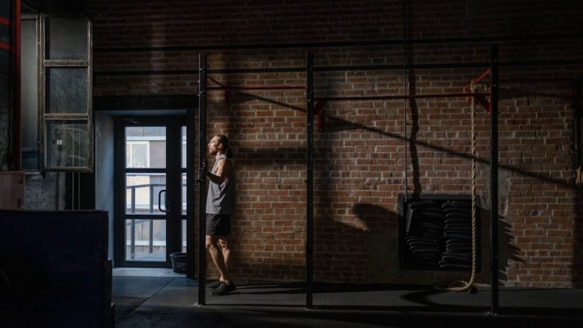 Muscular athlete standing pensively in gym, casting shadows on brick wall.